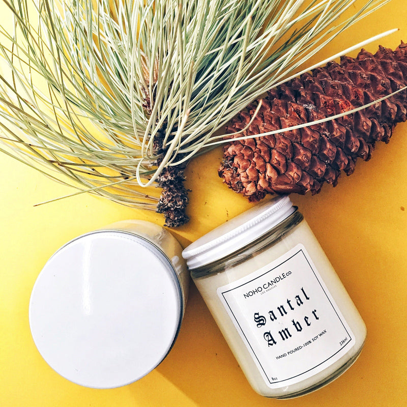 Two Santal Amber candle jars, one upright, and one on its side, next to a pinecone and pine needles for artistic flair.